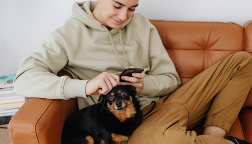 Young man sitting on couch with his little dog and scrolling on his cell