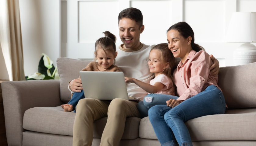 parents and two young daughters sitting on couch looking at laptop 