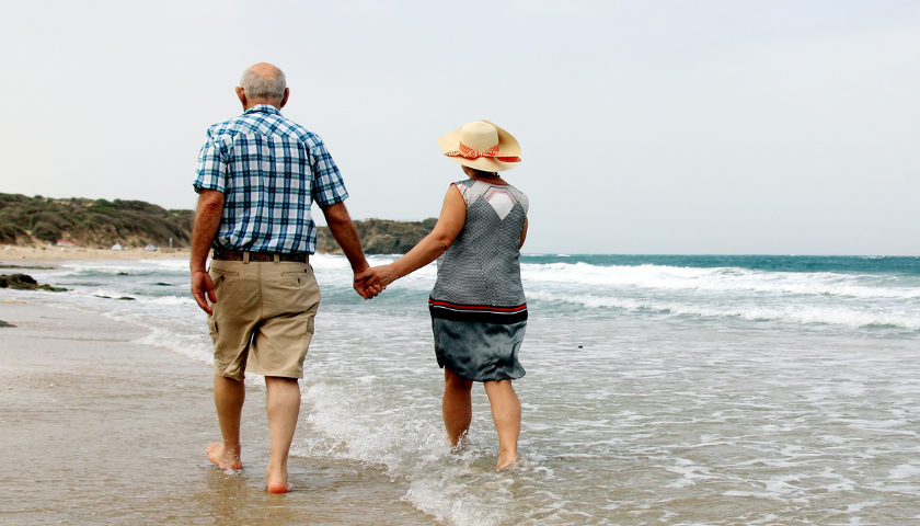 older couple holding hands walking on sandy beach next to ocean