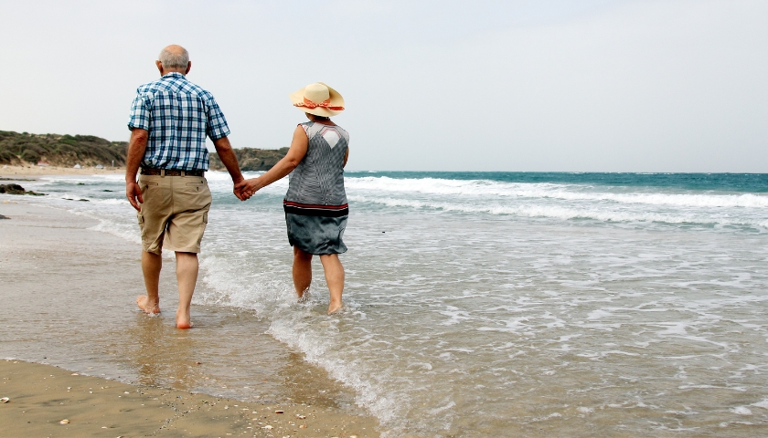 Elderly couple walking on beach holding hands.